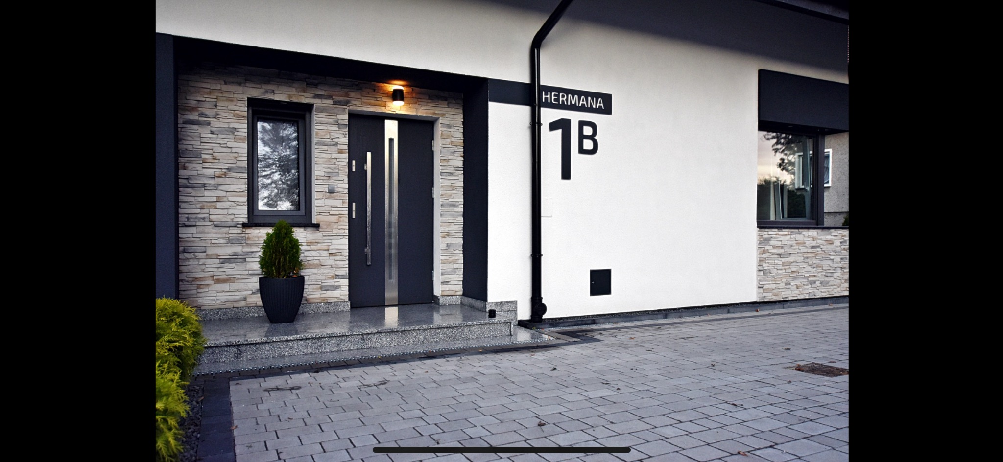 Nowoczesna elewacja domu z kamienną okładziną, antracytowymi oknami i drzwiami, oraz numerem budynku 'Herman 1B'.