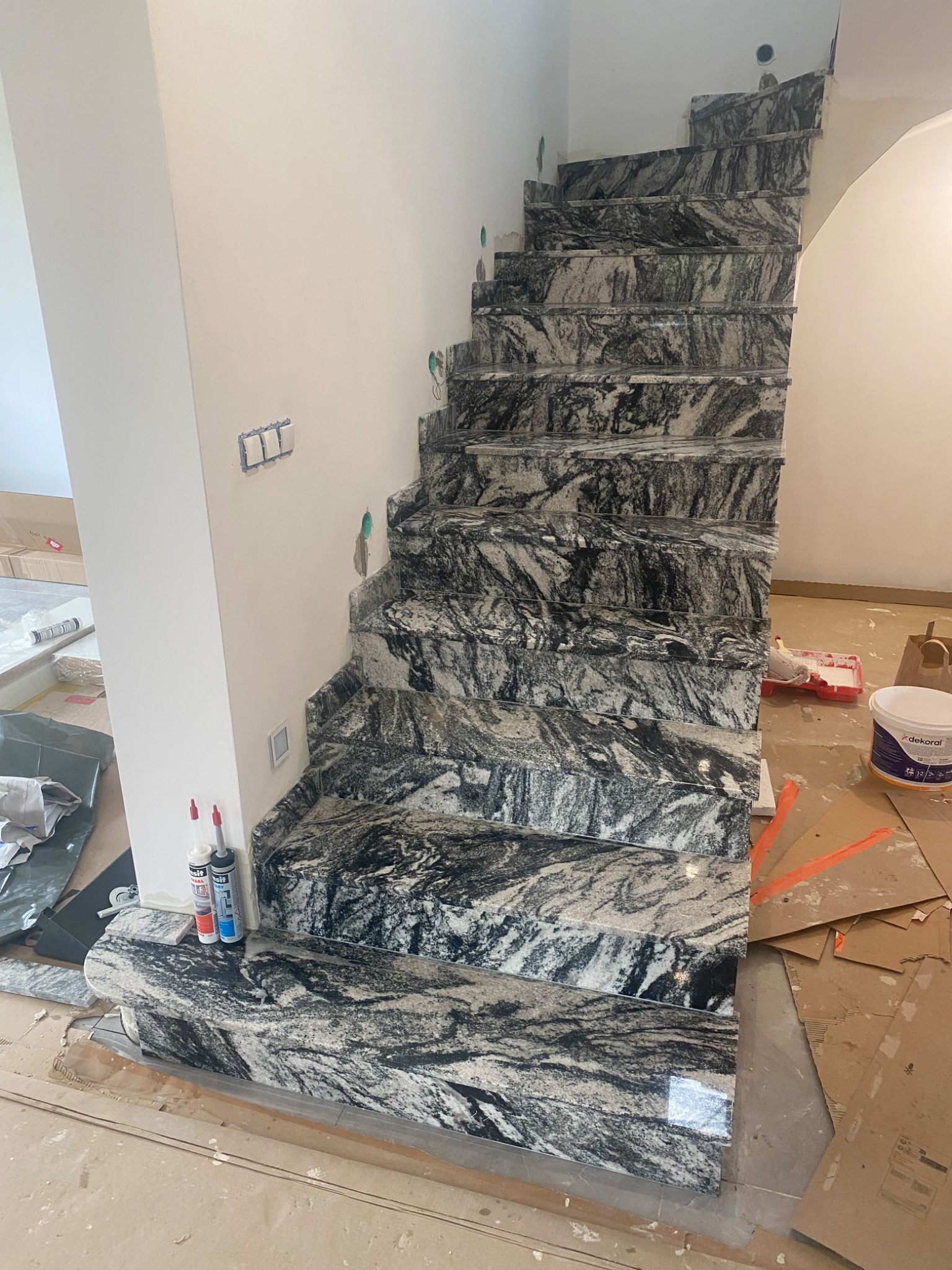 Schody wewnętrzne wyłożone marmurem o wzorze przypominającym fale, widoczne ślady prac wykończeniowych, silikon i puszka farby w tle.