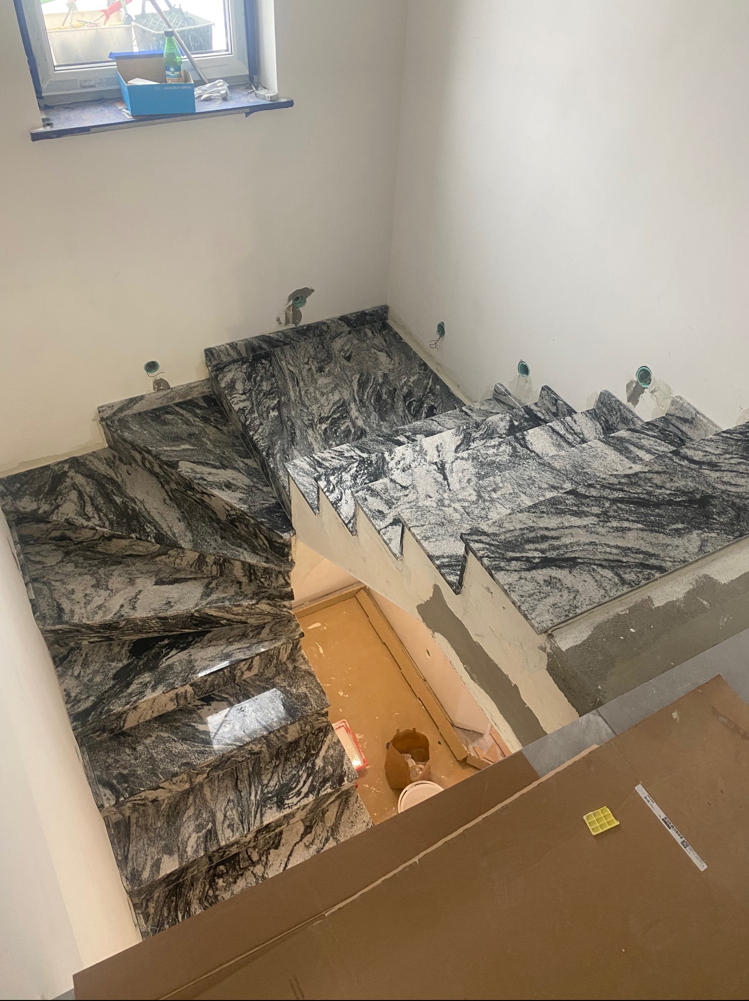 Schody wewnętrzne wyłożone płytami z szarego marmuru z wyraźnym, ciemnym żyłkowaniem, w trakcie montażu, widoczne niedoskonałości i zaprawa murarska, zabezpieczone podłogi.