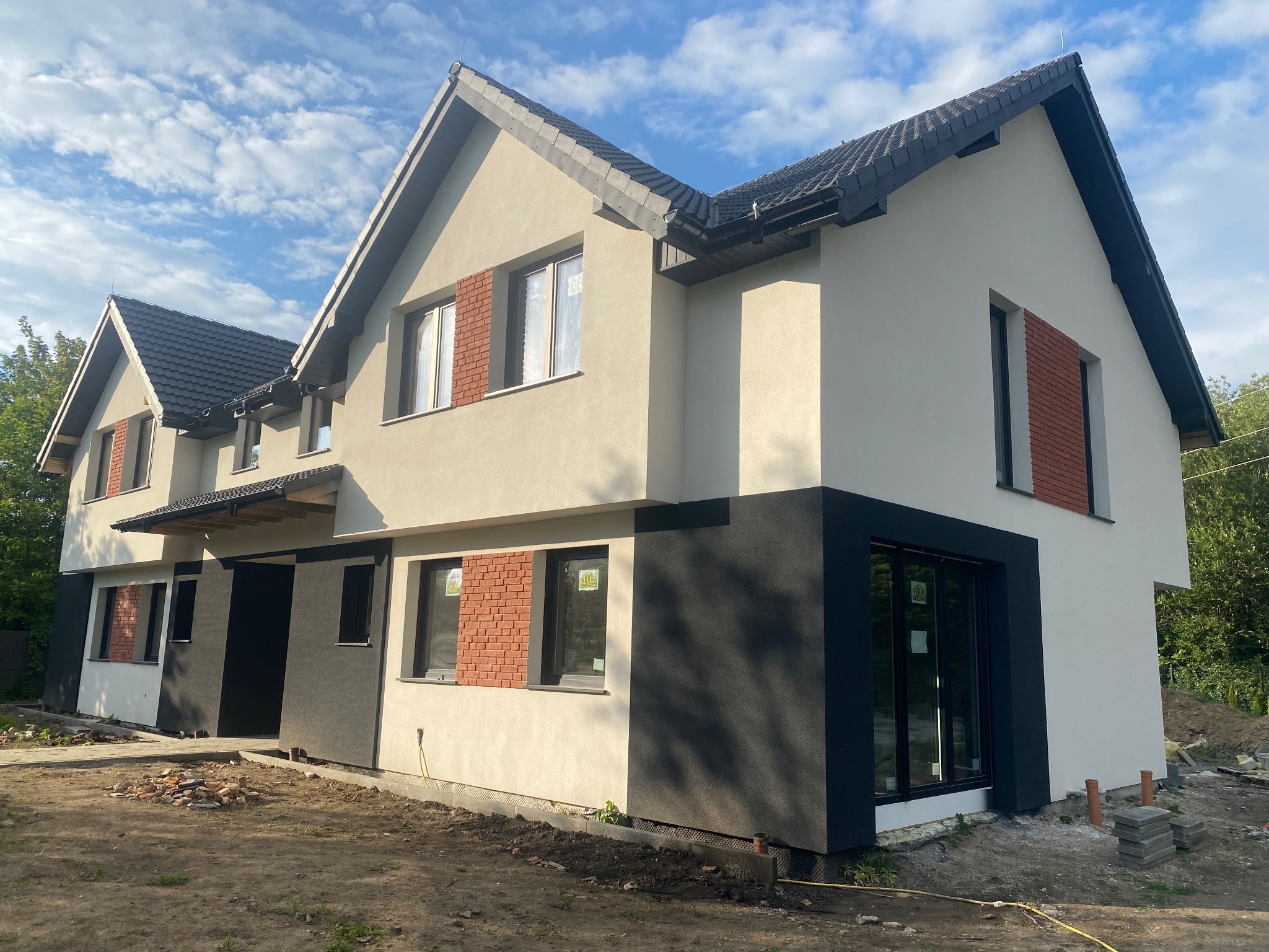 Nowoczesny dom dwukondygnacyjny w trakcie wykańczania elewacji, z fragmentami ścian w kolorze jasnego beżu i grafitu, okna z ceglaną dekoracją, czarny dach i częściowo zagospodarowany teren wokół.