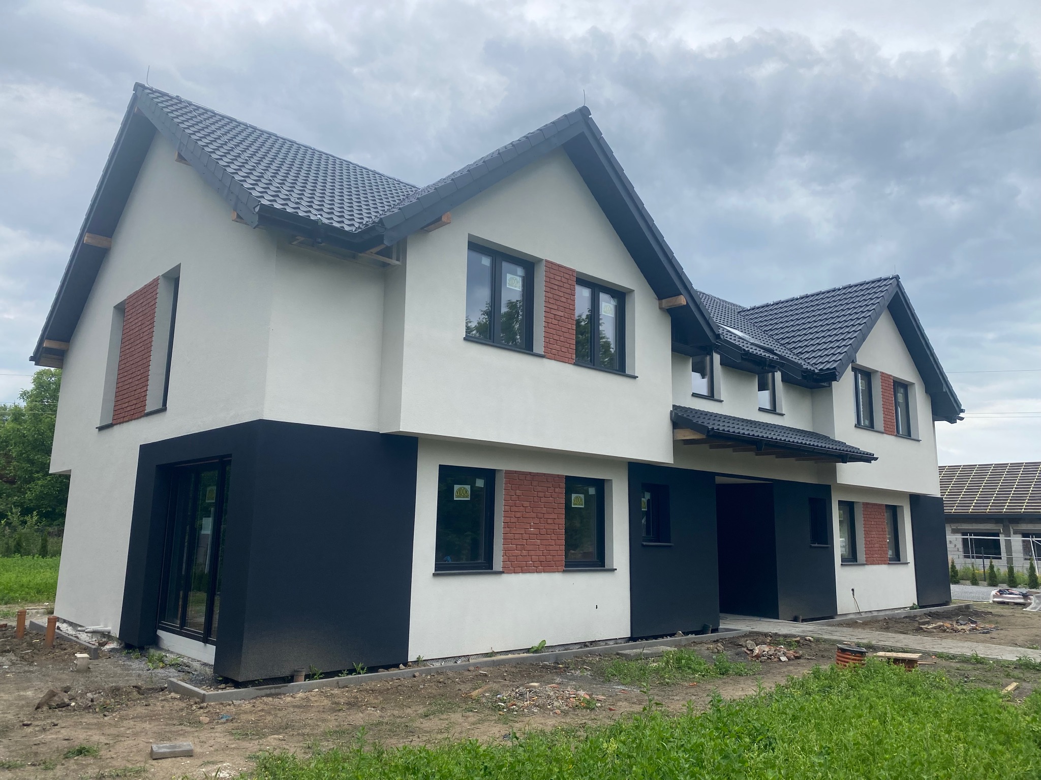 Dwukondygnacyjny dom w trakcie wykończenia elewacji: białe ściany z czarnym cokołem, fragmenty z cegły klinkierowej wokół okien, grafitowa dachówka karpiówka, teren wokół domu w trakcie prac ziemnych.