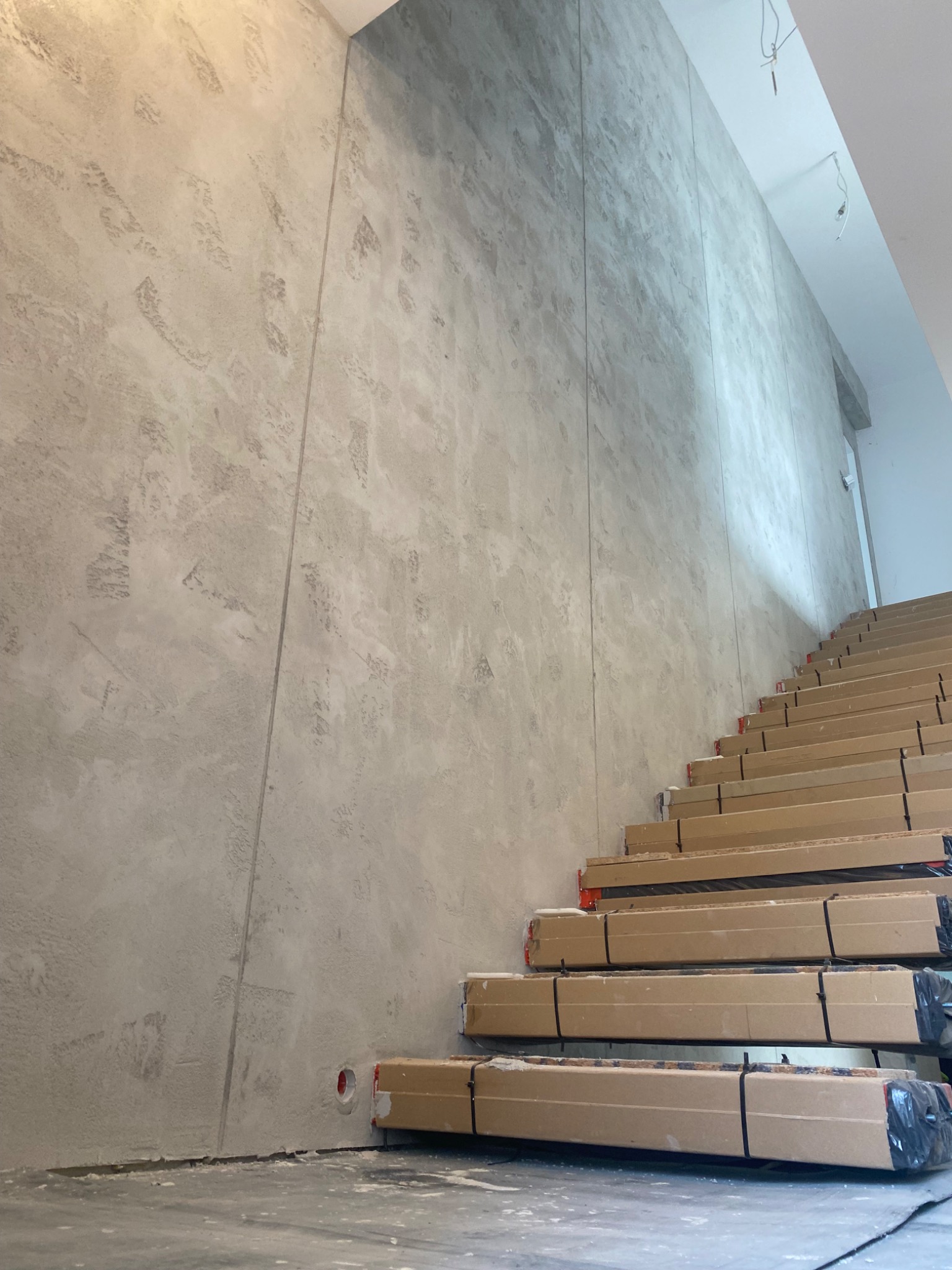 Ściana pokryta dekoracyjną szarą gładzią, z widocznymi pionowymi łączeniami paneli, obok schody zabezpieczone kartonem na czas prac wykończeniowych.