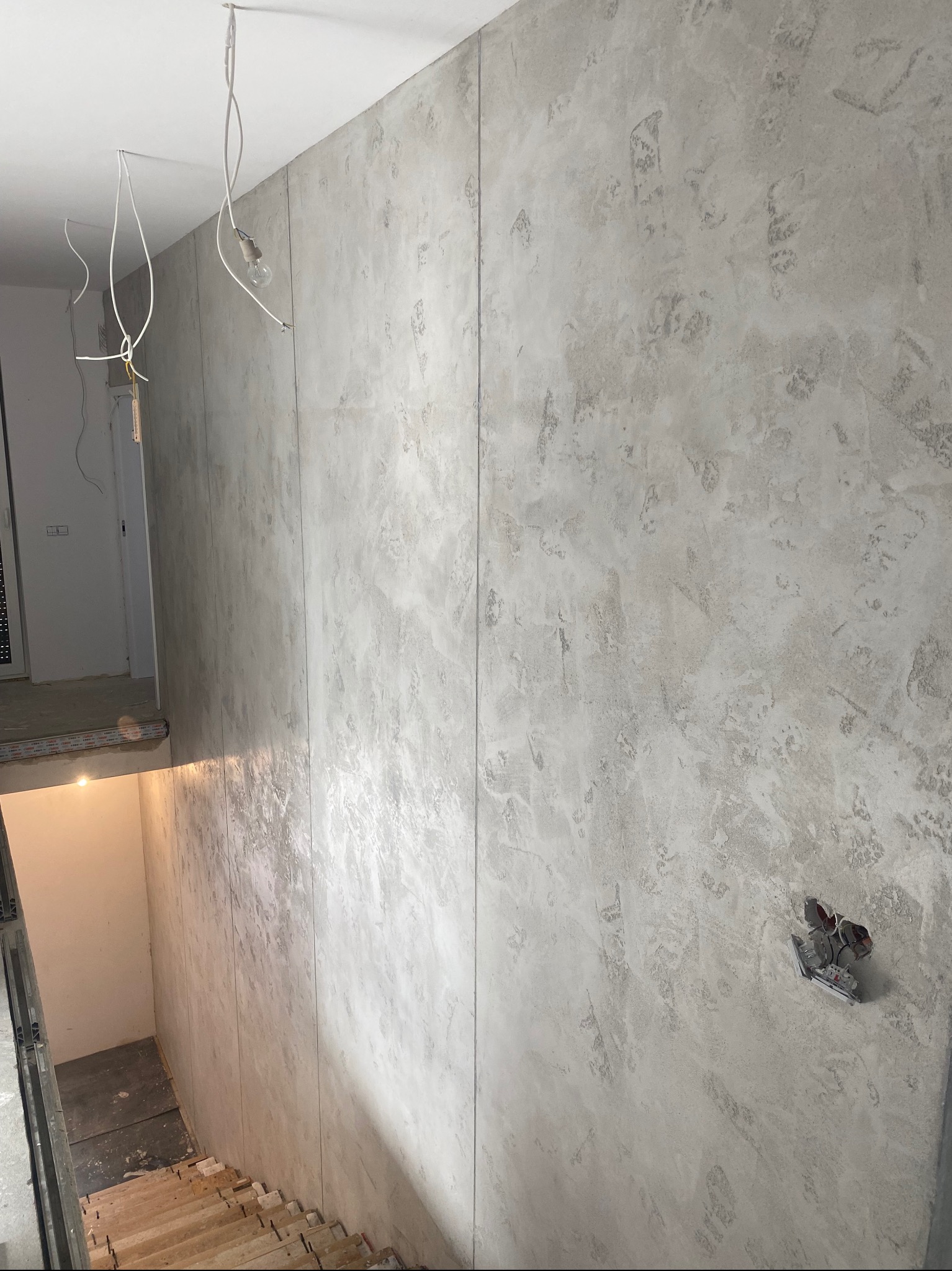 Ściana z efektem dekoracyjnym w odcieniach szarości, widoczne kable zwisające z sufitu i przygotowane gniazdko elektryczne, fragment schodów w tle