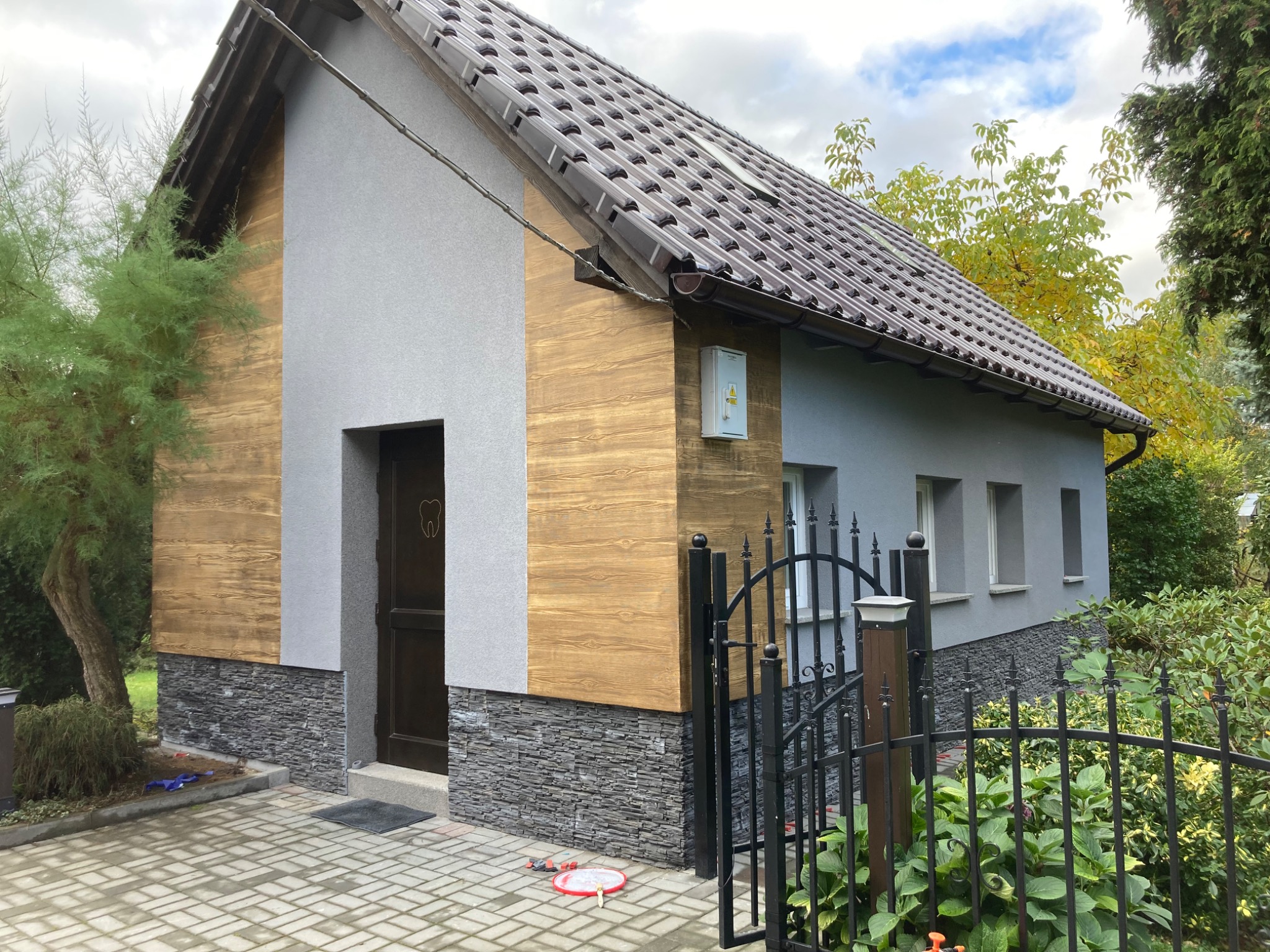 Elewacja budynku z elementami drewnopodobnymi i kamiennymi, szara ściana z oknami, brązowy dach, czarne ogrodzenie, kostka brukowa na podjeździe, zieleń wokół.