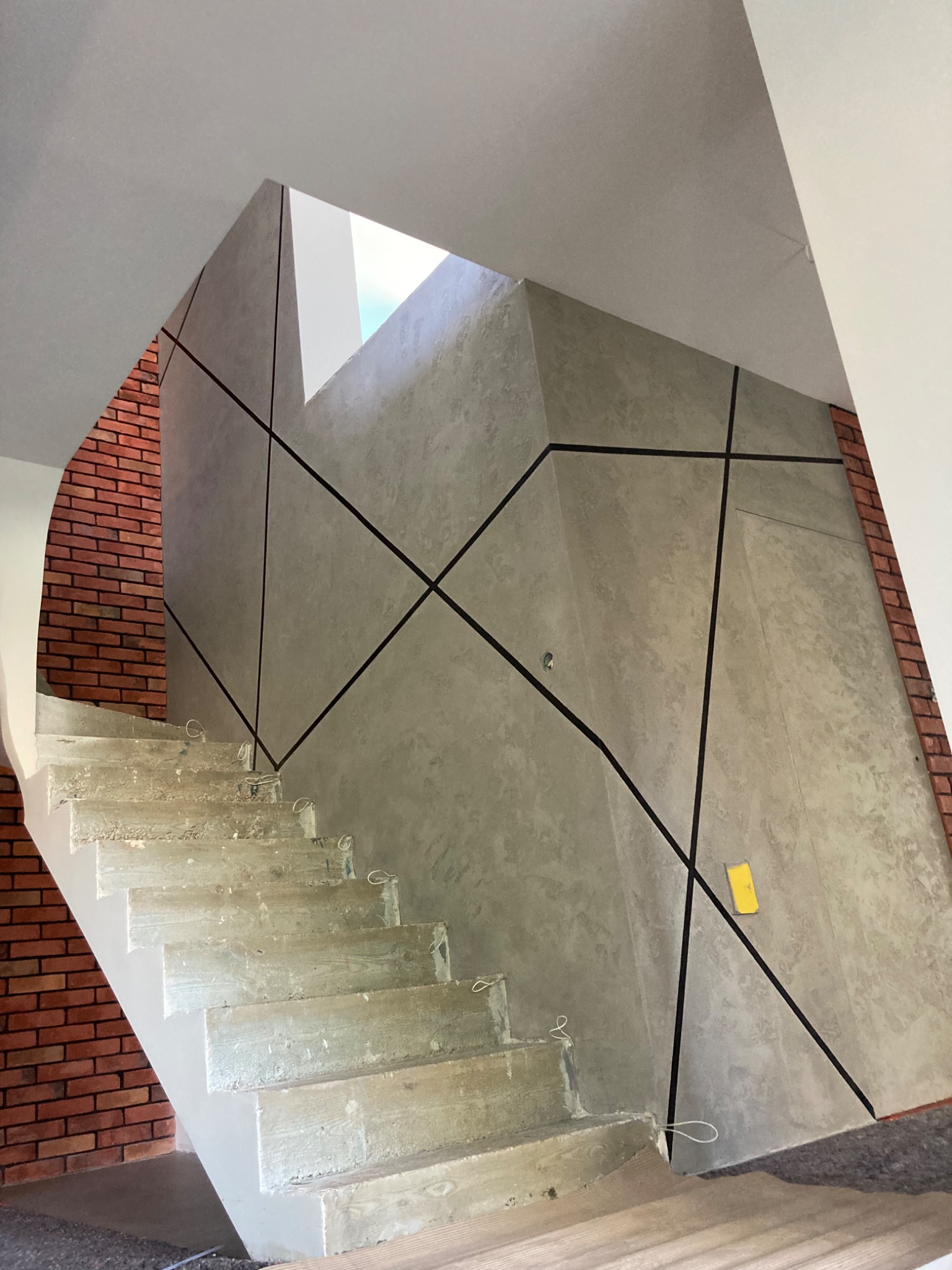 Klatka schodowa w trakcie wykańczania: surowe betonowe schody, ściana z dekoracyjną szarą strukturą i czarnymi liniami, fragment ściany z czerwonej cegły, widoczne przewody na schodach.