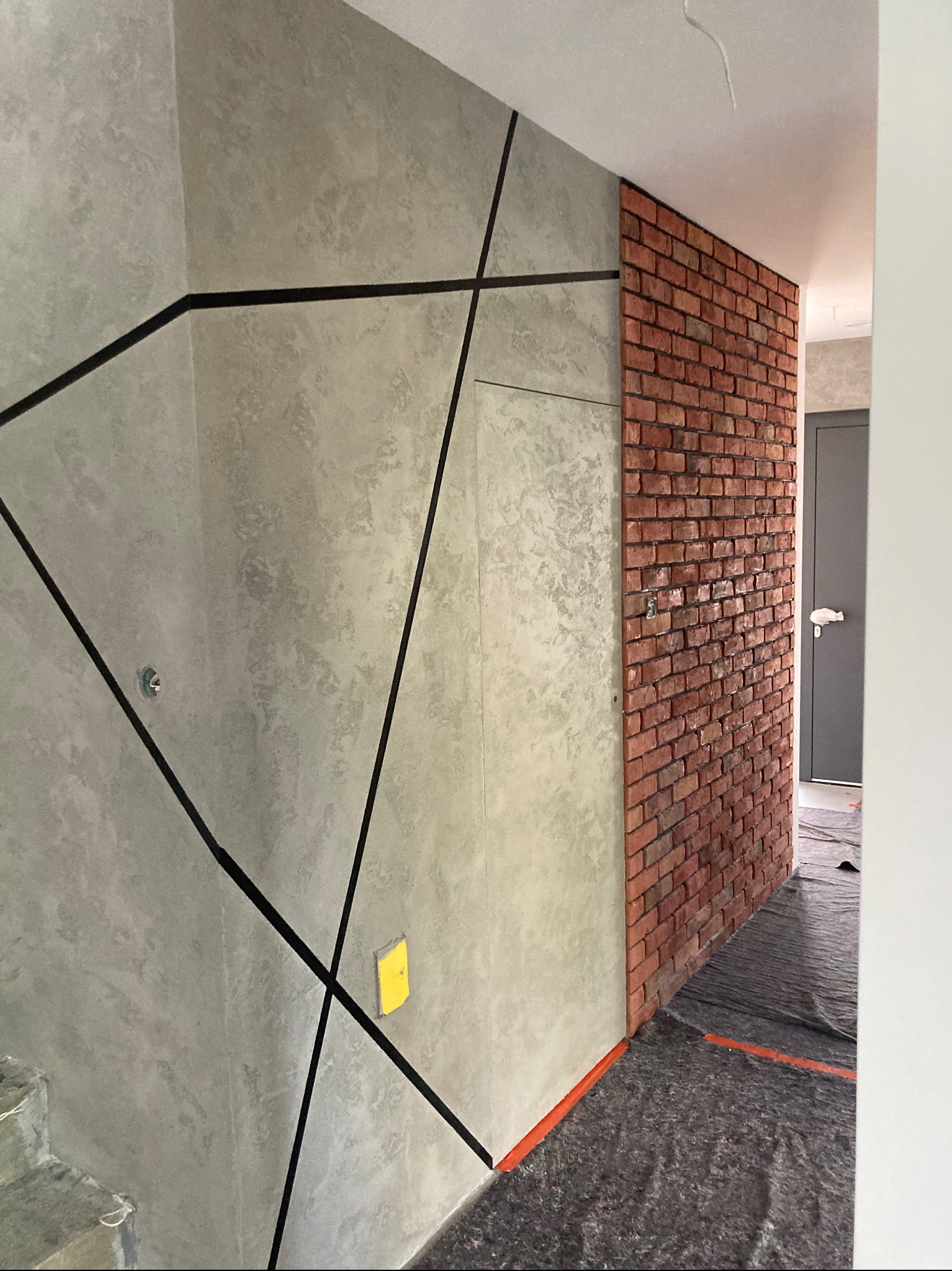 Ściana z dekoracyjną, szarą strukturą i czarnymi liniami tworzącymi geometryczny wzór, sąsiadująca z fragmentem ściany wyłożonej czerwoną cegłą, zabezpieczona podłoga folią ochronną.