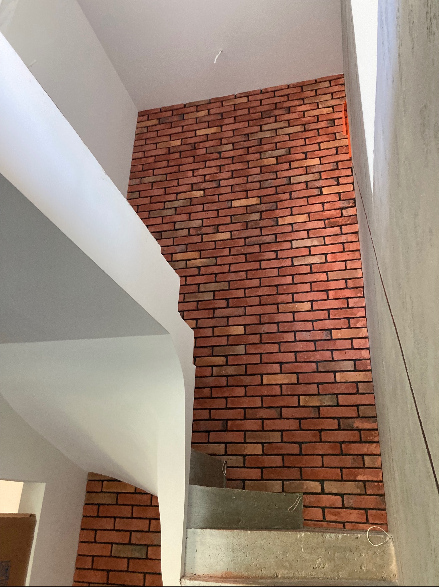 Ściana wykończona czerwoną cegłą klinkierową, widok ze schodów w surowym stanie, z elementami białej konstrukcji stropu i ściany w tle.
