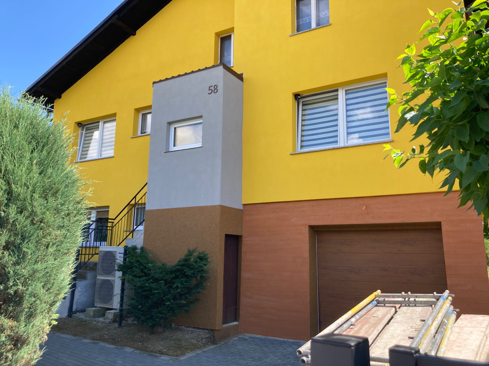 Żółty dom dwukondygnacyjny z szarym pionowym elementem z numerem 58, brązową dolną częścią i garażem, widoczne rusztowanie i zieleń.