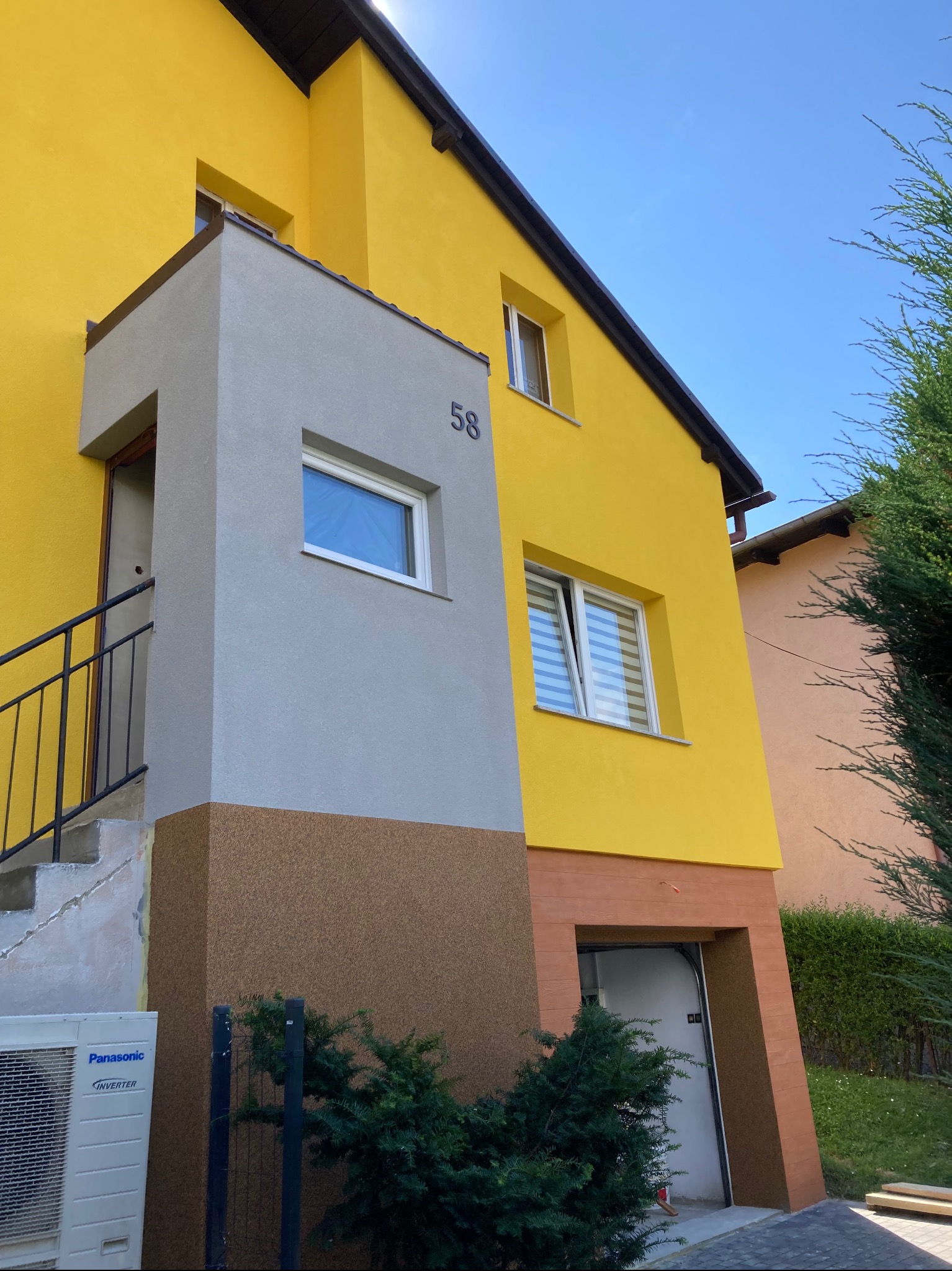 Dwukondygnacyjny dom z żółtą elewacją i szarym gankiem z numerem 58, widoczny klimatyzator Panasonic, brązowe akcenty elewacyjne i zieleń w tle.