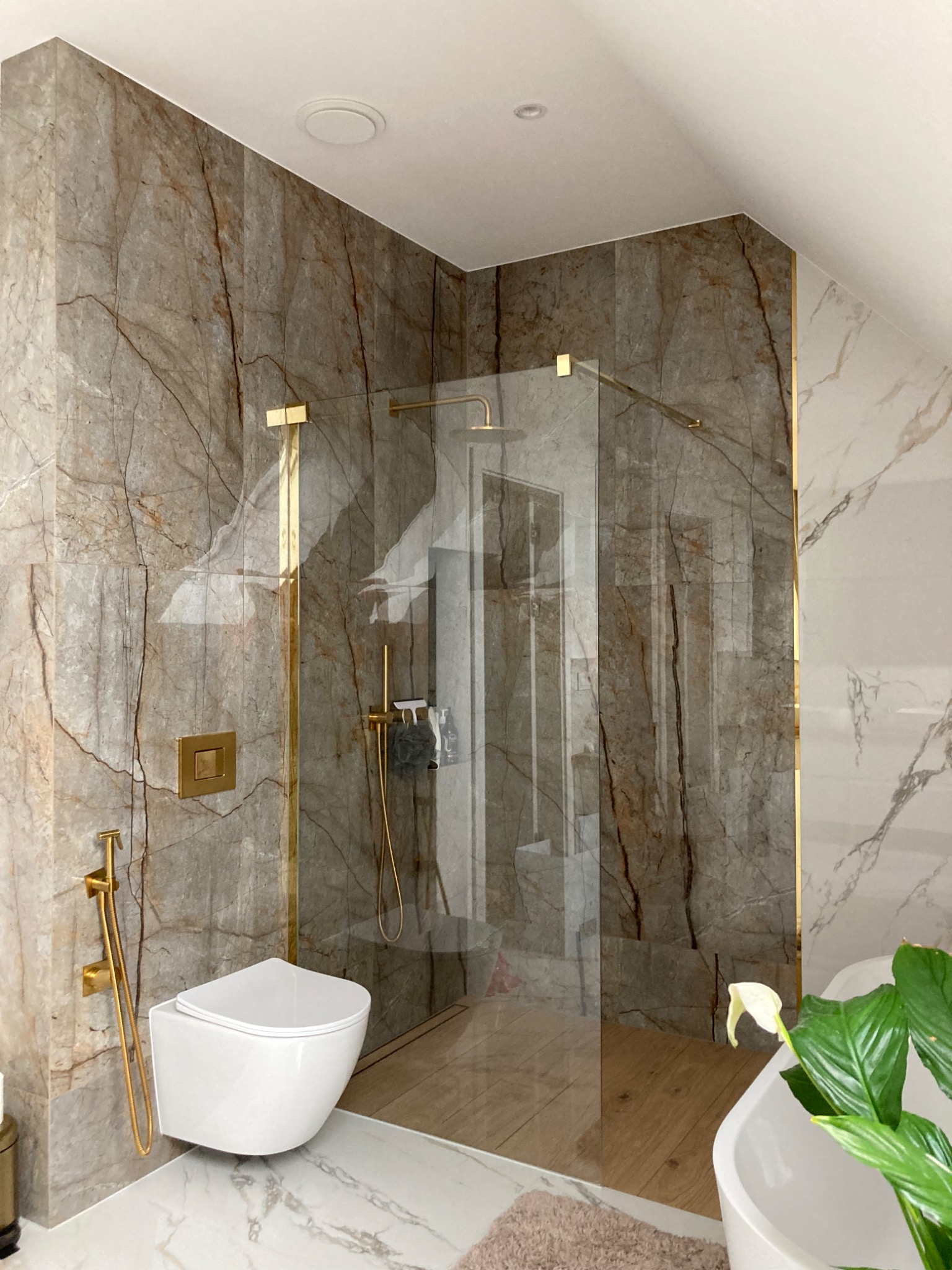 Nowoczesna łazienka w odcieniach beżu i bieli z marmurowymi płytkami, złotymi akcentami armatury i kabiną prysznicową typu walk-in, wisząca toaleta z bidetką i subtelny akcent roślinny.