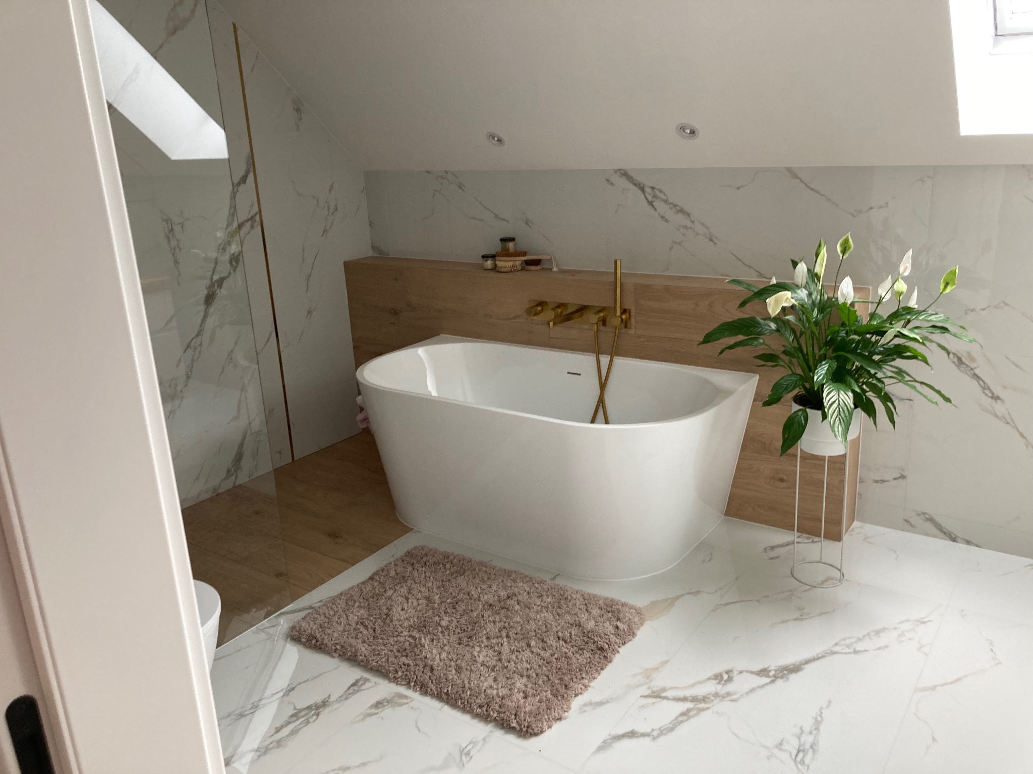 Nowoczesna łazienka na poddaszu z wolnostojącą białą wanną, złotą armaturą, marmurowymi płytkami i rośliną doniczkową.