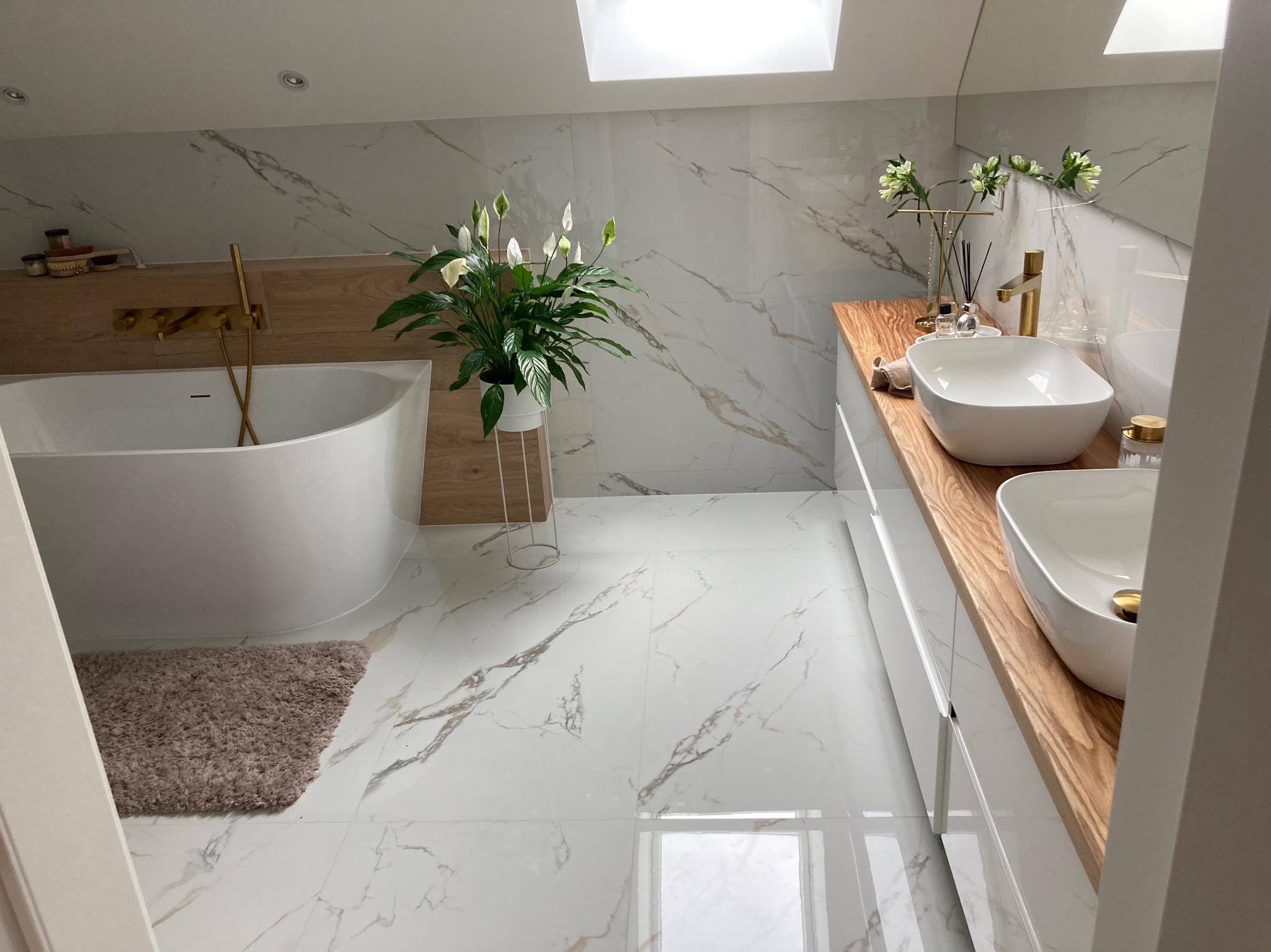 Jasna łazienka na poddaszu z marmurowymi płytkami, wolnostojącą wanną z złotą armaturą, umywalkami nablatowymi na drewnianym blacie i roślinami w doniczkach.