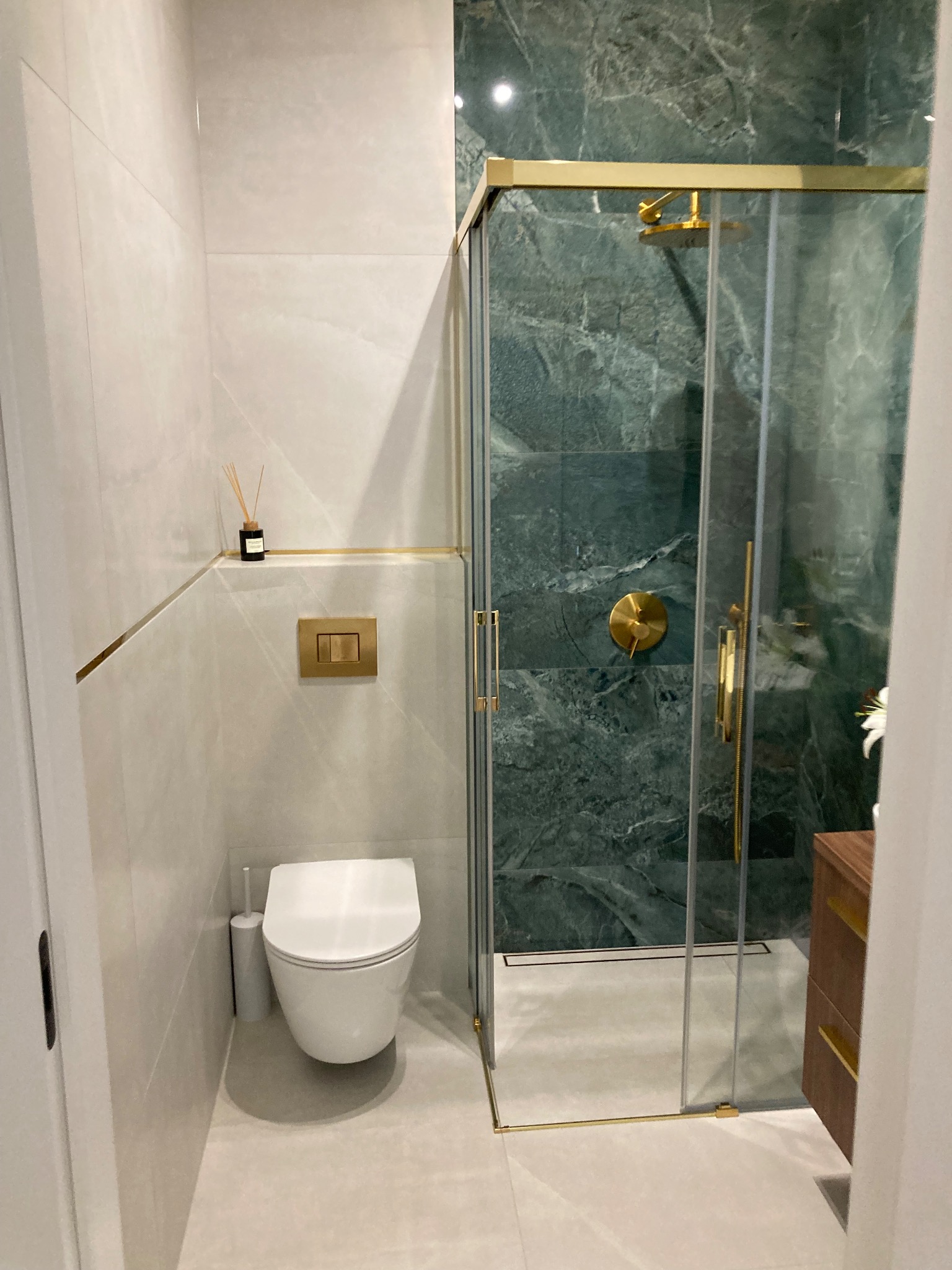 Nowoczesna łazienka z kabiną prysznicową typu walk-in z zielonym marmurem, złotymi detalami, podwieszaną toaletą i subtelnymi akcentami zapachowymi.