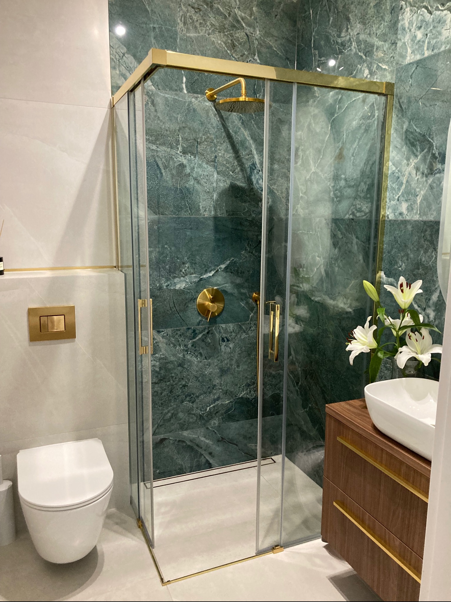 Nowoczesna łazienka z kabiną prysznicową w złotej ramie, marmurowymi ścianami w odcieniach zieleni, wiszącą toaletą i umywalką nablatową z kwiatami.