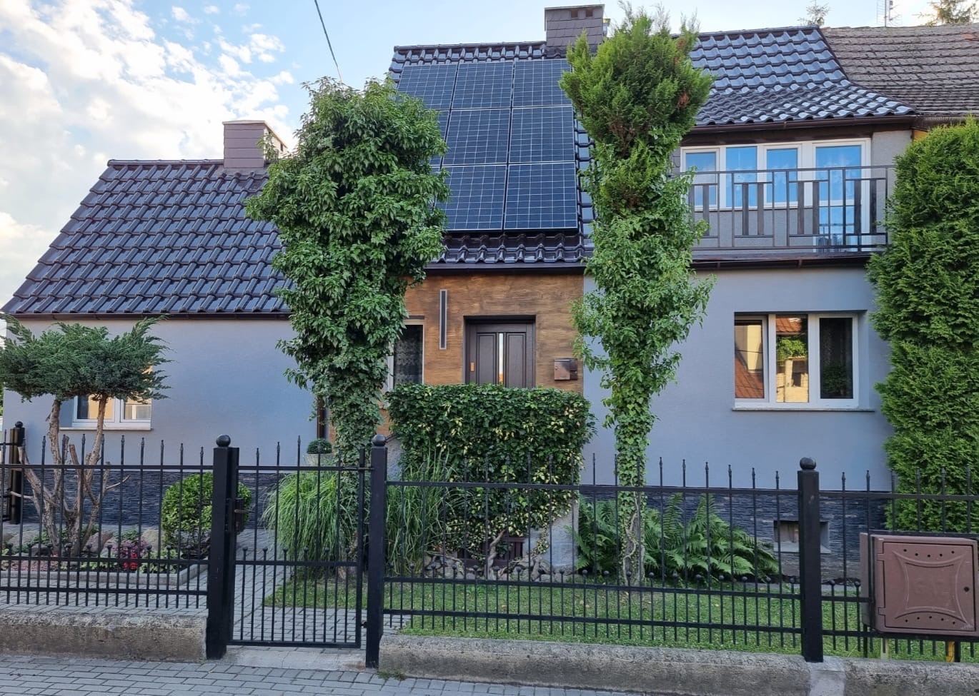 Dwukondygnacyjny dom z szarą elewacją, brązową wstawką i panelami słonecznymi na dachu, widoczny zza czarnego metalowego ogrodzenia i zieleni.
