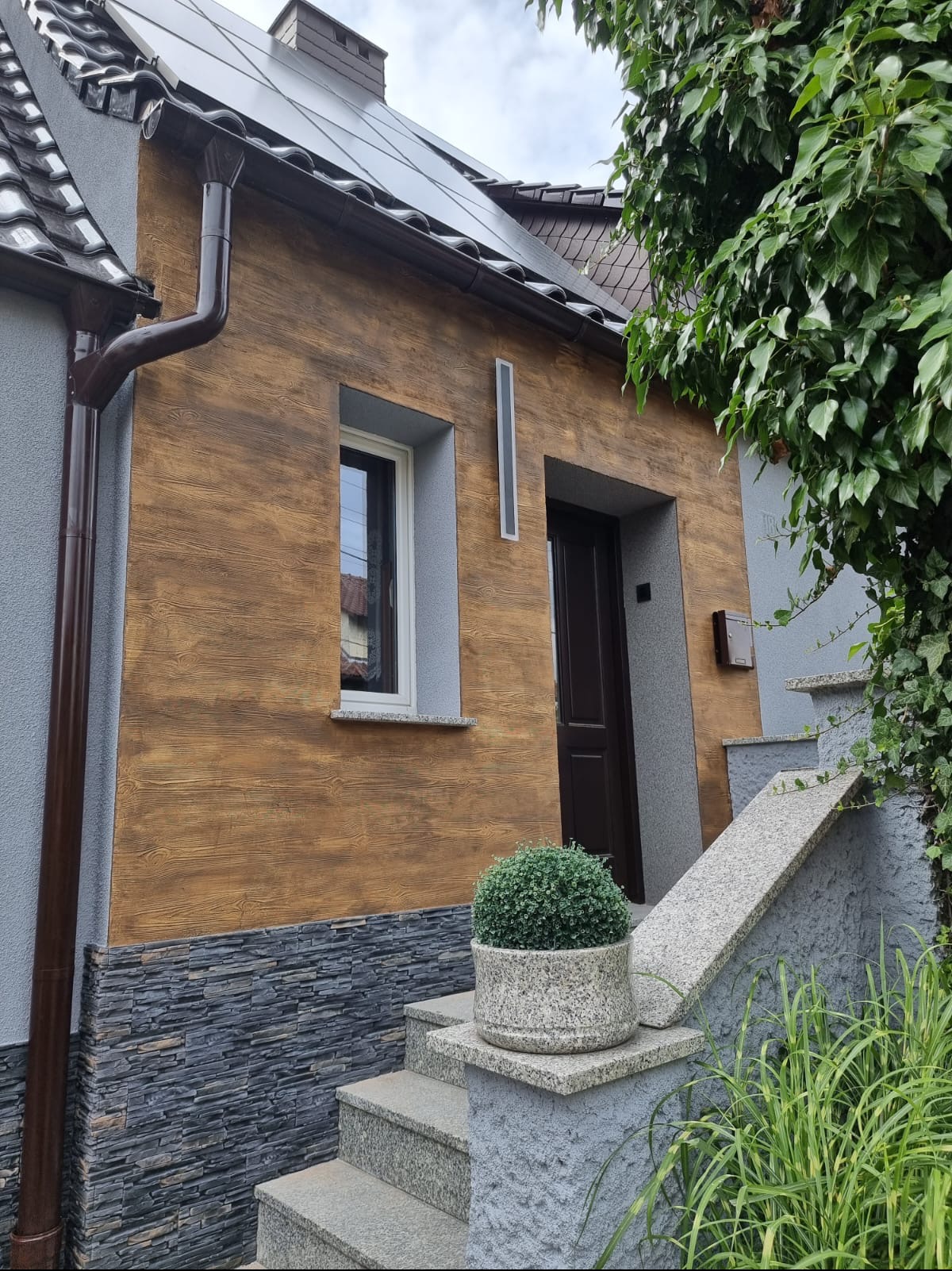 Jasnobrązowa elewacja domu z fakturą drewna, szara podmurówka z kamienia łupanego, brązowa rynna i granitowe schody wejściowe