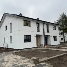 Bliźniak w nowoczesnym stylu z białymi ścianami i czarnymi oknami, świeżo wykończony, z przygotowanym terenem pod trawnik i kostką brukową.