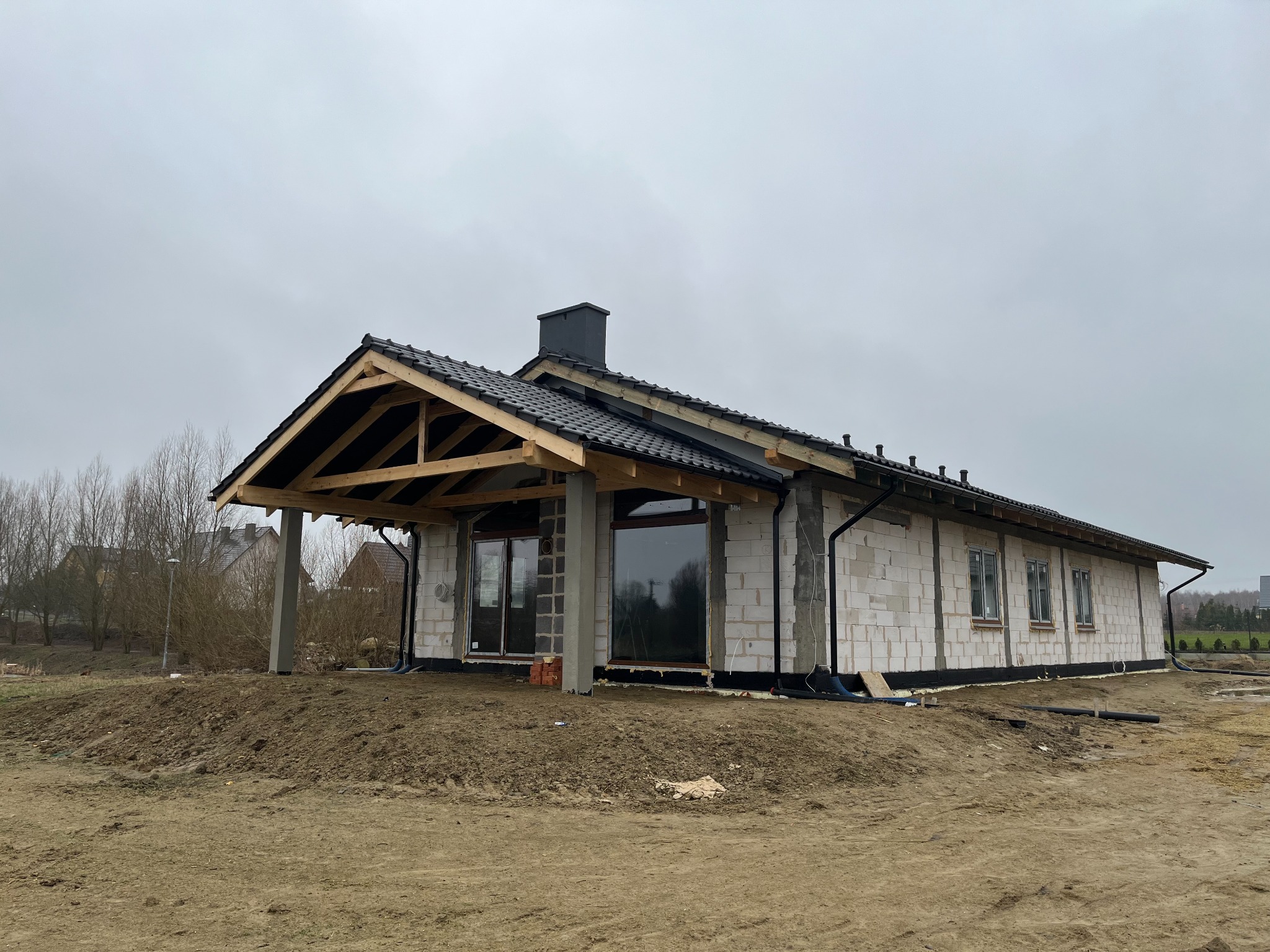 Surowy stan domu z białego bloczka, widoczny dach z czarną dachówką, brak elewacji, teren wokół w stanie ziemnym, widoczne okna i drzwi balkonowe.