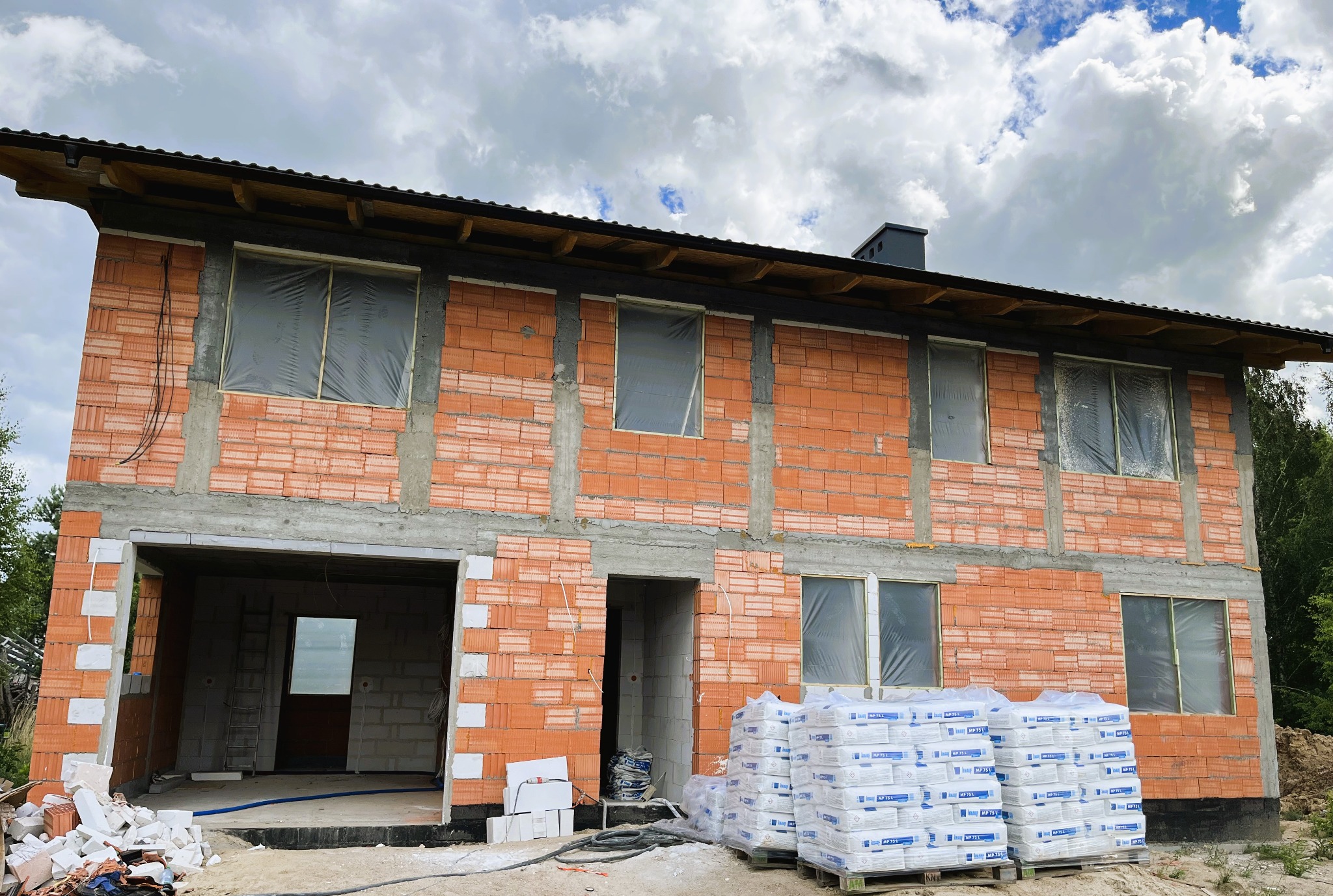 Dwukondygnacyjny budynek w trakcie budowy z czerwonej cegły, z oknami zabezpieczonymi folią, palety z zaprawą murarską na pierwszym planie, pochmurne niebo w tle.