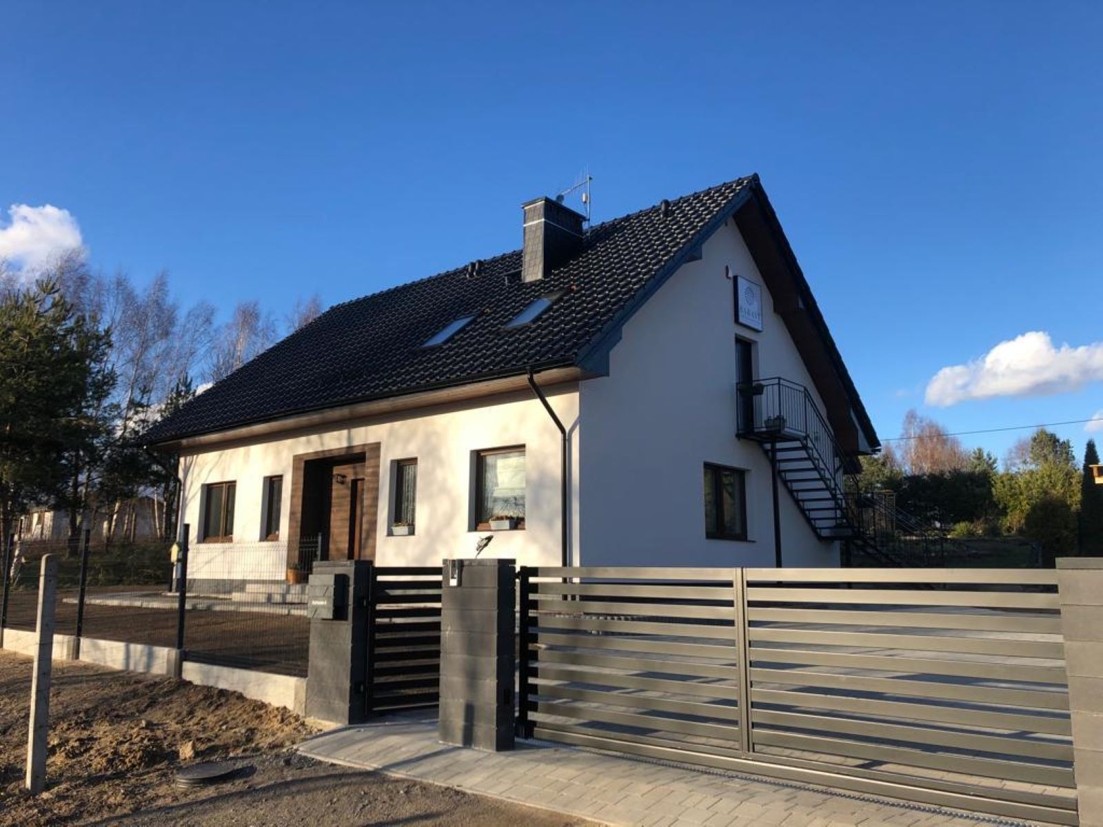 Nowoczesny dom jednorodzinny z czarną dachówką, zewnętrznymi schodami stalowymi i przesuwną bramą wjazdową z szarych paneli, otoczony ogrodzeniem i świeżo wyrównanym terenem.