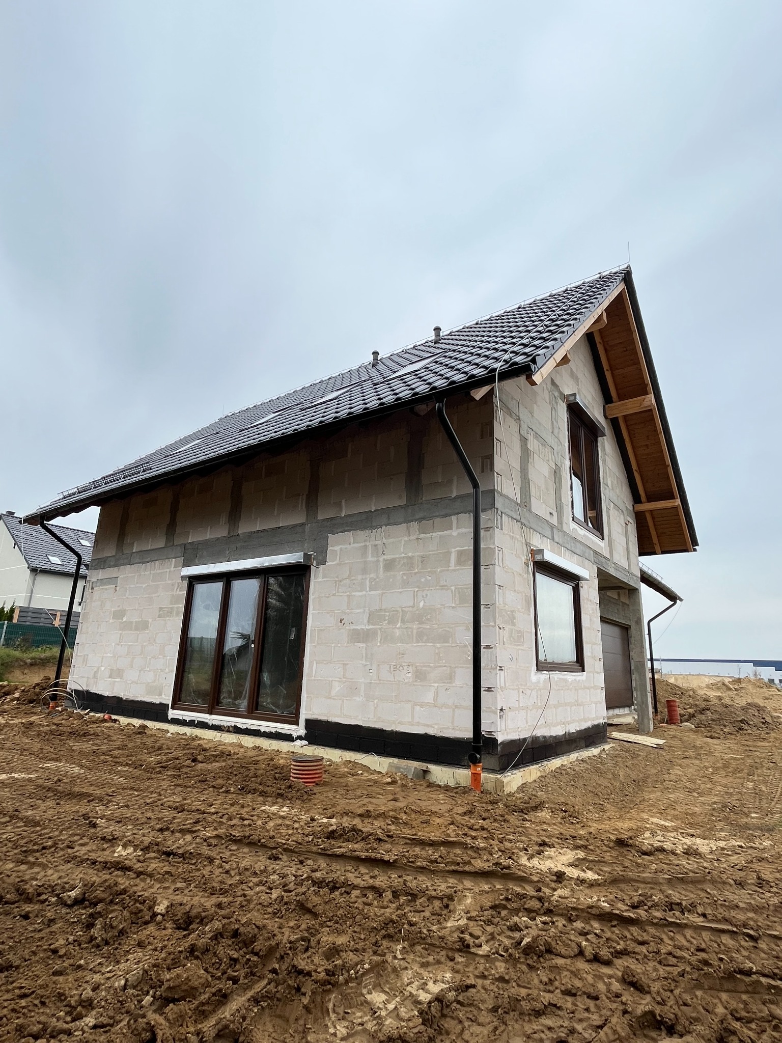 Surowy stan domu z białego pustaka z ciemną dachówką, widoczne okna z brązowymi ramami, teren wokół domu pokryty błotem.