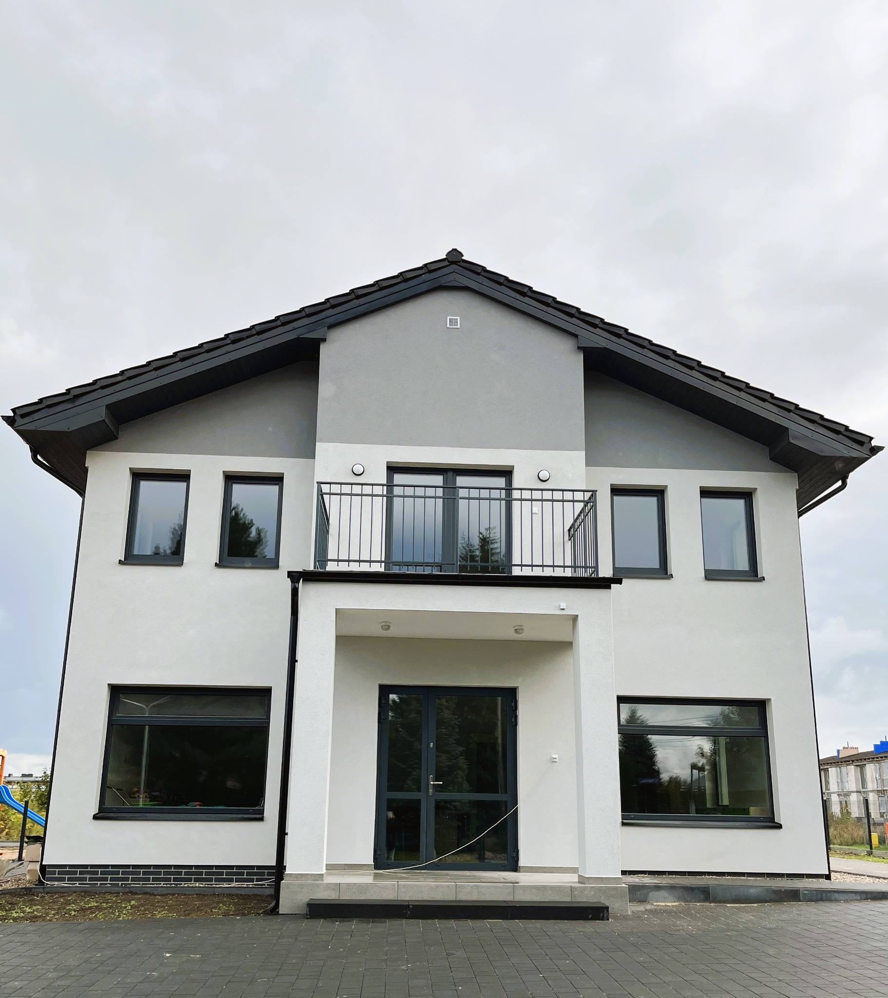 Nowoczesny dwukondygnacyjny dom z balkonem, wykończony w bieli i szarości, z ciemną stolarką okienną i dachówką, widok z dołu na tle pochmurnego nieba.