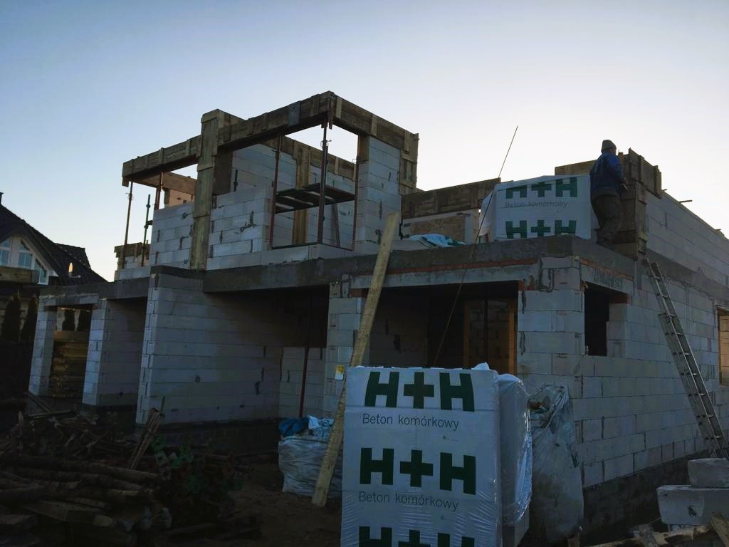 Budowa domu jednorodzinnego z betonu komórkowego H+H w trakcie wznoszenia ścian, widoczny pracownik na dachu i palety z materiałem budowlanym.