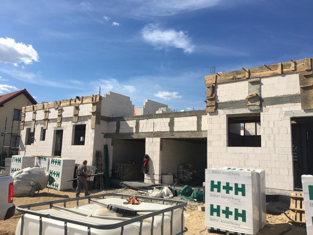 Budowa domu z betonu komórkowego H+H na etapie wznoszenia ścian, widoczne szalunki na nadprożach, pracownik w tle, palety z bloczkami i zbiornik na wodę na pierwszym planie.