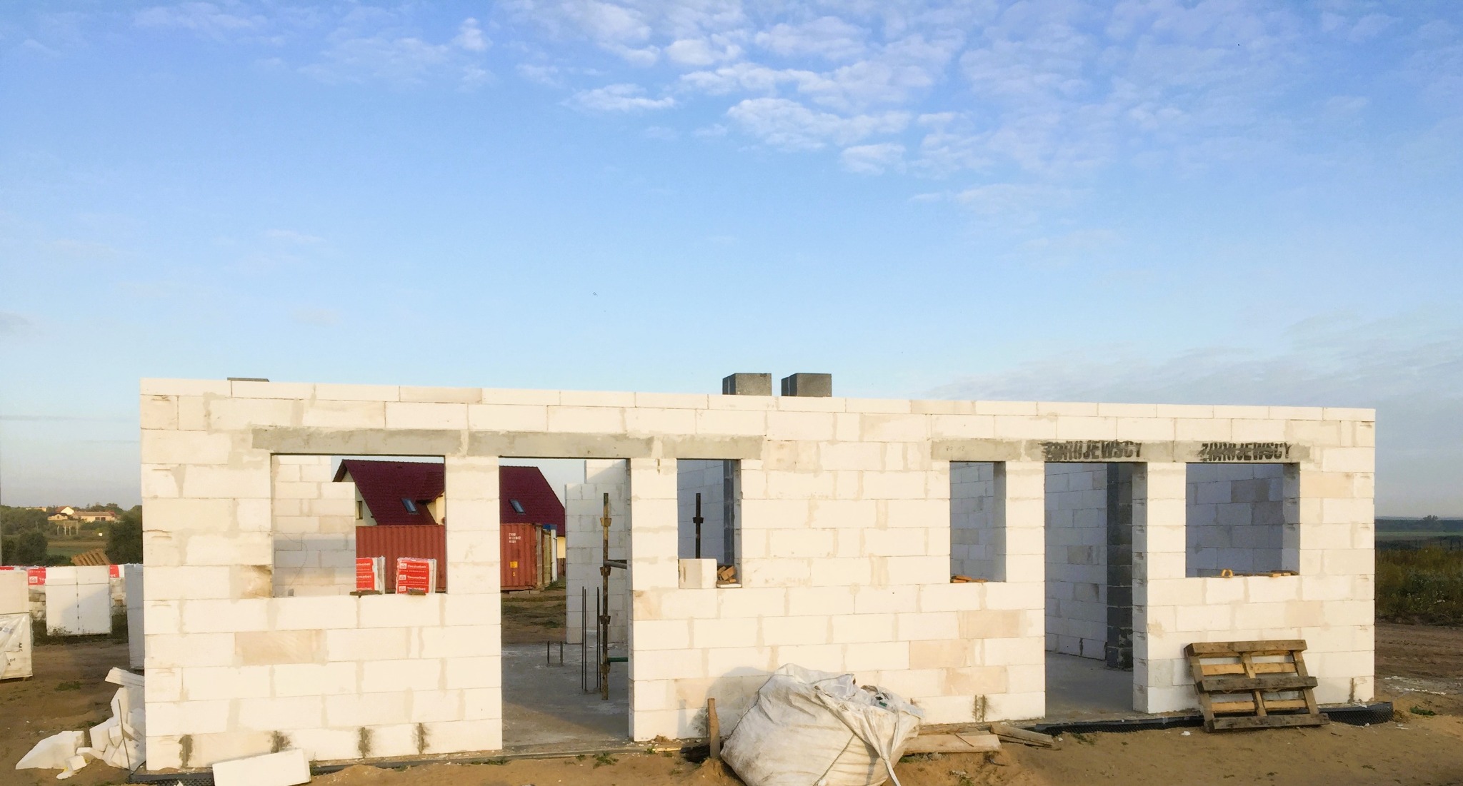 Wzniesione ściany parterowego budynku z bloczków betonu komórkowego, widoczne otwory okienne i drzwiowe, w tle częściowo widoczny czerwony dach innego budynku i błękitne niebo.