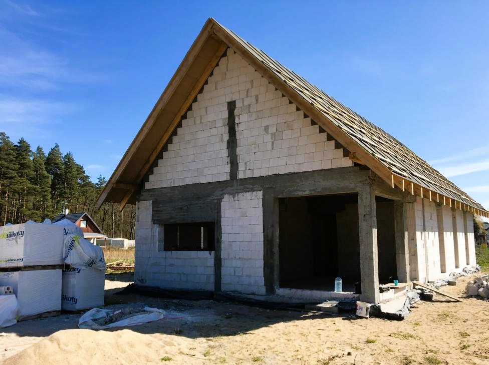 Surowy stan budynku z białych bloczków, widoczne niezadaszone okno i otwory drzwiowe, częściowo wykonana więźba dachowa, na pierwszym planie palety z bloczkami Silikaty.