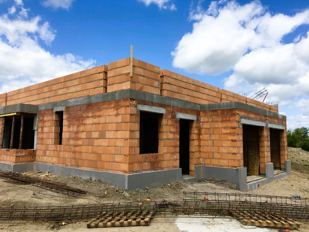 Surowy stan domu z czerwonej cegły w Pruszczu Gdańskim, widoczne zbrojenia i fundamenty, niebo z chmurami w tle.