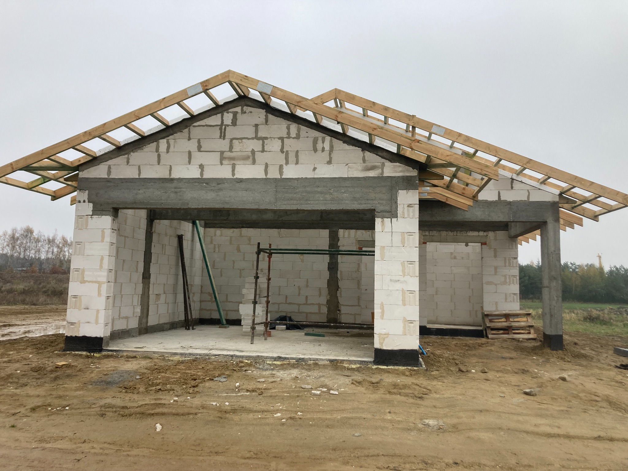 Surowy stan domu murowanego z betonu komórkowego z widoczną konstrukcją dachu z drewna, fundamenty zabezpieczone izolacją przeciwwilgociową, teren wokół w stanie niezagospodarowanym.
