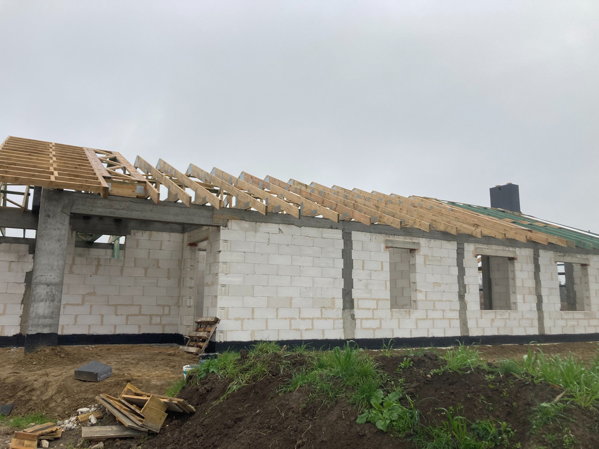 Surowy stan domu z białych bloczków, widoczne zbrojenia i drewniana konstrukcja dachu, fragment izolacji przeciwwilgociowej przy ziemi, na pierwszym planie gruz budowlany i zieleń.