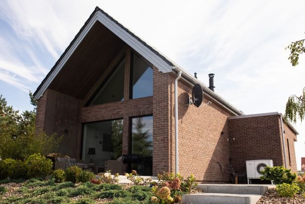 Nowoczesny dom z cegły klinkierowej z dużymi oknami, anteną satelitarną na ścianie i pompą ciepła na zewnątrz.