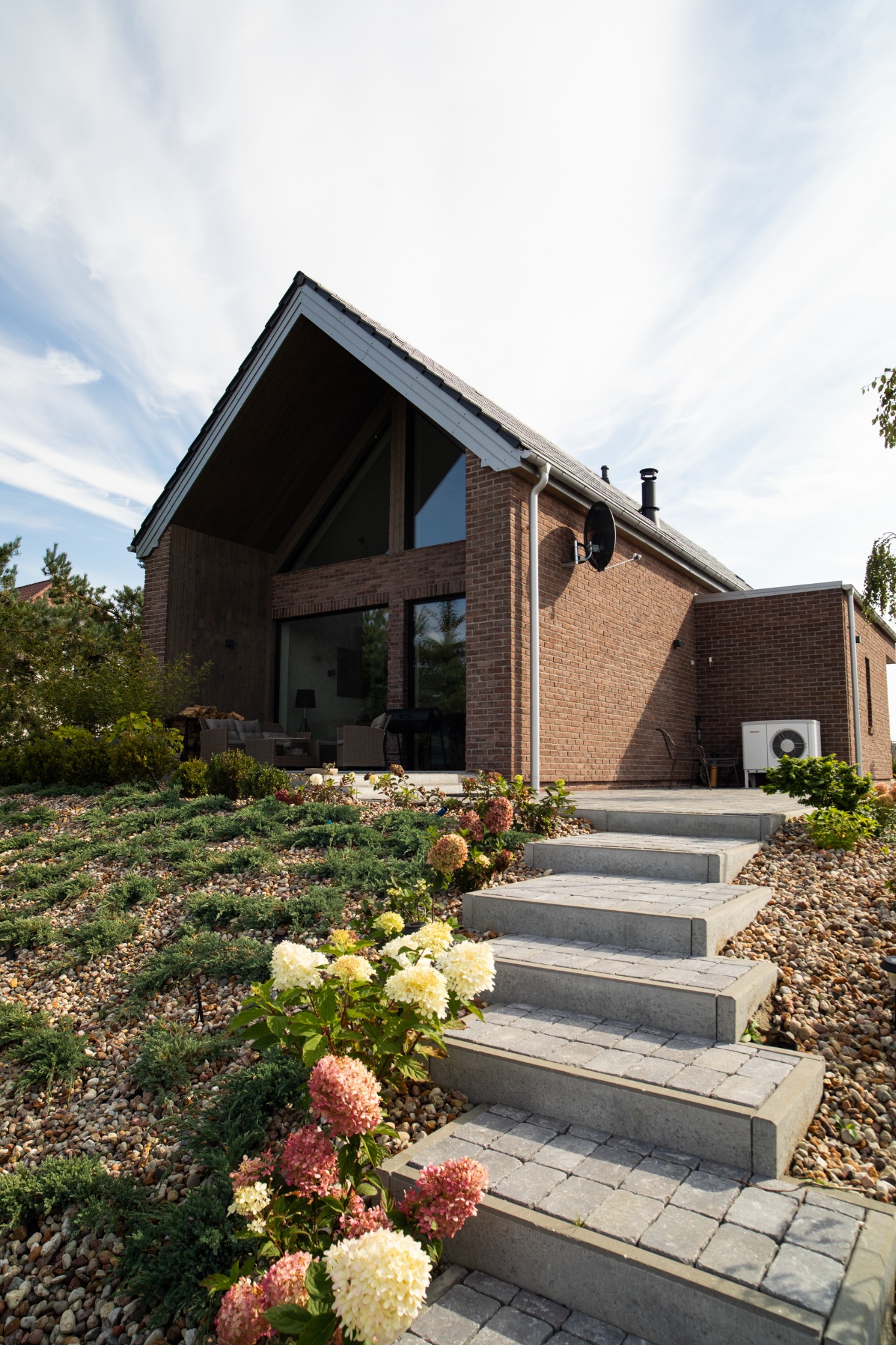 Nowoczesny dom z czerwonej cegły z dużymi oknami i schodami prowadzącymi do wejścia, otoczony ogrodem z kamieniami i kwitnącymi hortensjami.