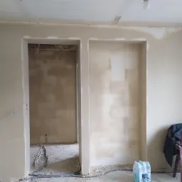 Wnętrze w trakcie remontu: ściany po nałożeniu tynku, widoczne otwory drzwiowe i wnęka, niezagruntowane fragmenty, przewody elektryczne wystające ze ściany, butelki wody i krzesło z torbą w rogu...