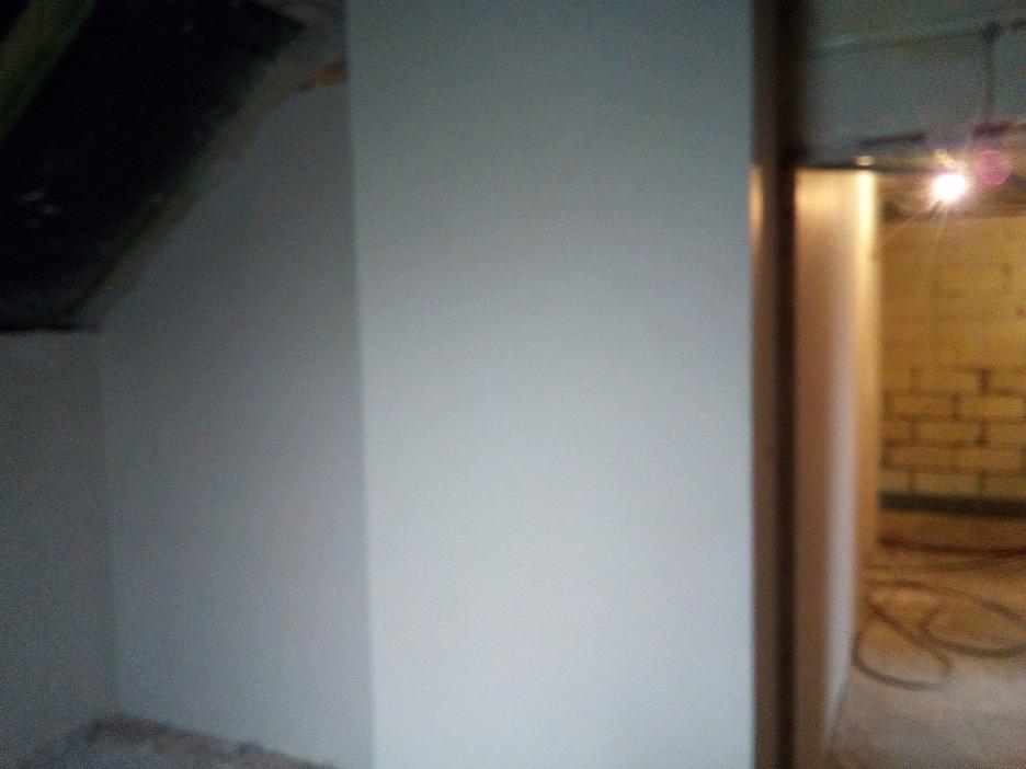 Świeżo otynkowana ściana w piwnicy, widoczna surowa konstrukcja stropu i fragment muru z cegły w przejściu do sąsiedniego pomieszczenia.