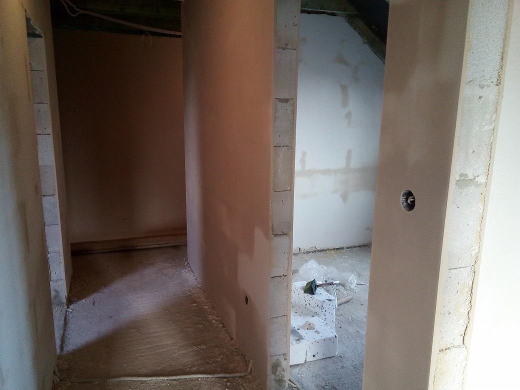 Surowe wnętrze w trakcie prac wykończeniowych: ściany pokryte świeżym tynkiem, widoczne bloczki betonowe i elementy instalacji elektrycznej, podłoga z resztkami materiałów budowlanych. W tle...