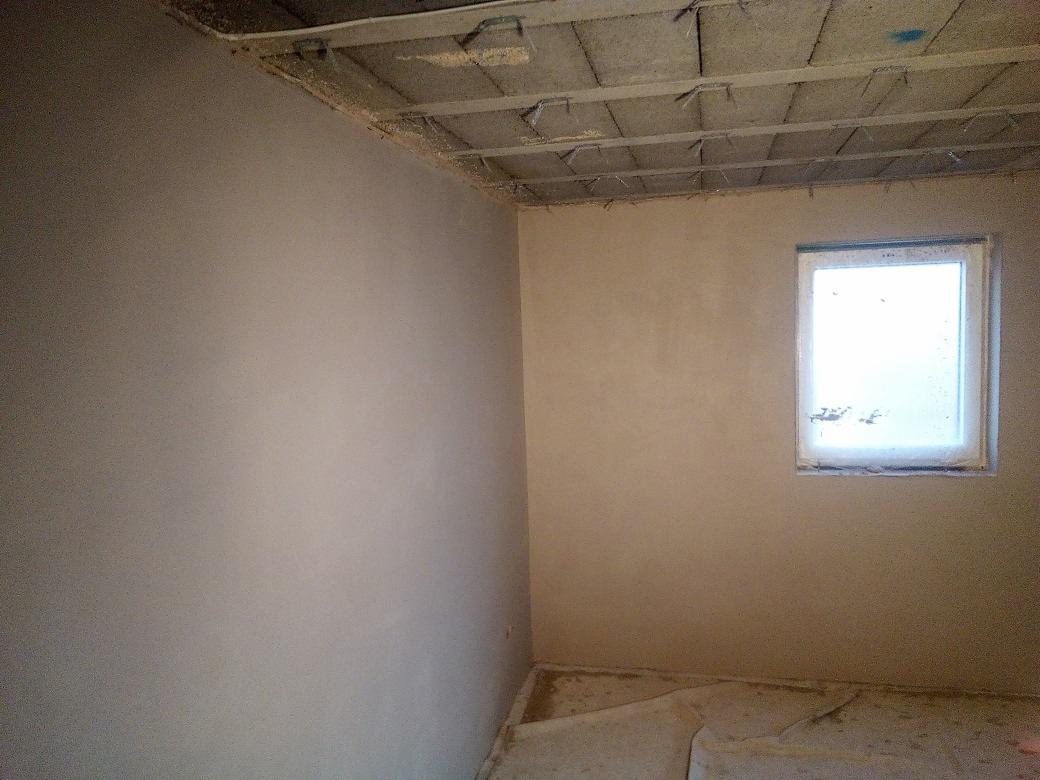 Surowe, szare tynki maszynowe na ścianach pokoju z małym oknem, widoczne ślady po pracach budowlanych na suficie i podłodze.