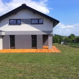 Nowoczesny dom z drewnianym tarasem, świeżo założonym trawnikiem i rabatą kwiatową wzdłuż ogrodzenia, widok w słoneczny dzień.