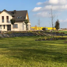 Nowoczesny dom jednorodzinny z zadbanym trawnikiem, świeżo posadzone drzewa wzdłuż podjazdu, w tle żółty budynek magazynowy.