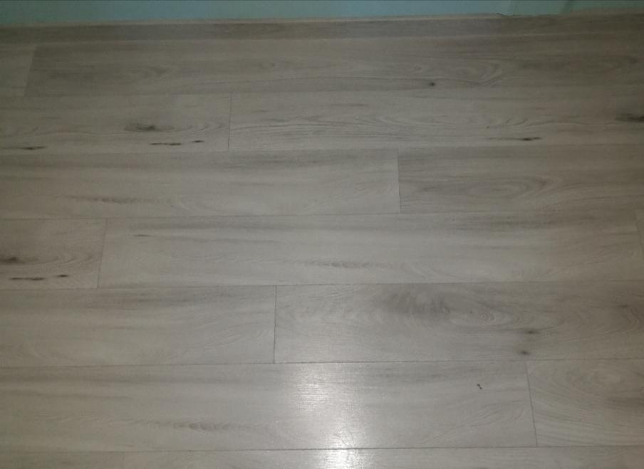 Jasne, szare panele podłogowe ułożone wzdłużnie, imitujące drewno z widoczną strukturą słojów i delikatnymi, ciemniejszymi plamkami, przy listwie przypodłogowej w odcieniu błękitu.