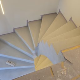 D2Team - Spiralne schody po nałożeniu gładzi, zabezpieczone taśmą malarską. Widoczna szpachelka i oznaczenia wymiarów na podłodze. Ujęcie z góry, oświetlenie ledowe.