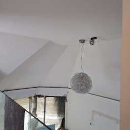 De-Jot - Wnętrze w trakcie wykończenia: białe ściany, sufit ze skosami, nowoczesna lampa wisząca, widoczne narzędzia i materiały budowlane. Ujęcie z góry, zza szklanej balustrady.