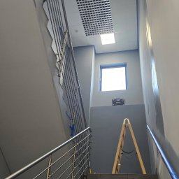 De-Jot - Malowanie wysokiej ściany klatki schodowej w budynku, z pracownikiem używającym wałka na długim kiju. Widoczne szare ściany, niebieska taśma maskująca, drabina i okno.