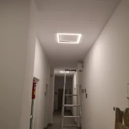 Korytarz biurowy po świeżym malowaniu na biało, widoczna aluminiowa drabina rozstawiona na środku, sufit kasetonowy z kwadratowymi lampami LED, podłoga w szarą kratę, w tle gaśnica wnękowa.