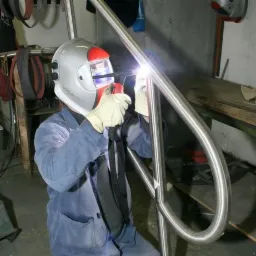 Spawanie poręczy ze stali nierdzewnej przez spawacza w masce ochronnej, widoczny błysk spawania i szlifowane spawy.