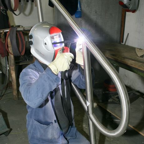 Spawanie poręczy ze stali nierdzewnej przez spawacza w masce ochronnej, widoczny błysk spawania i szlifowane spawy.