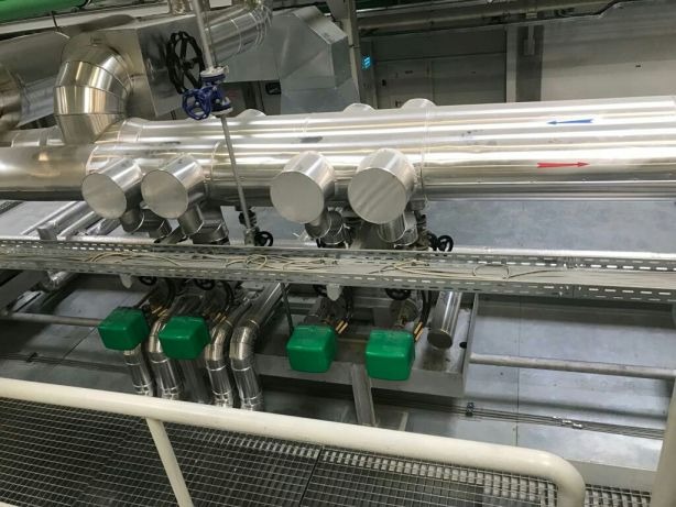 Instalacja przemysłowa z rurami w izolacji, zaworami i zielonymi osłonami, widoczne strzałki wskazujące kierunek przepływu.