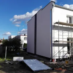 Boczny widok ocieplonego, białego budynku z rusztowaniem, metalową drabiną i rynną, na tle błękitnego nieba z chmurami, z widocznymi elementami ocieplenia na ziemi.